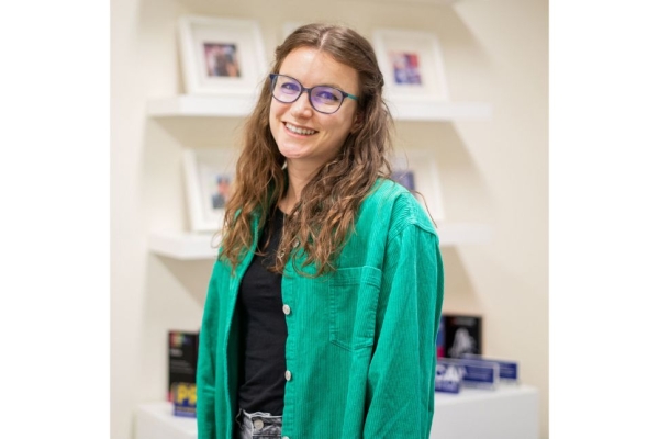 A portrait of Leah Elston-Thompson, a white woman with long brown hair wearing glasses and a green shirt over a black top stands in front of shelving